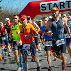 Zawodnicy biegną pod czerwoną bramką startową. Wszyscy przestrzegają zasad triathlonu, skupieni na rywalizacji.