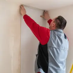 Mann tapeziert eine Wand in einer Wohnung. Er hält eine Tapetenbahn an die Decke, um sie anzubringen.