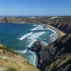 Malownicze wybrzeże Portugalii z klifami, piaszczystą plażą i turkusowym oceanem. Białe fale rozbijają się o brzeg, a w oddali widać białe domki.