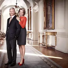 Królowa Belgii, uśmiechnięta, stoi obok króla w eleganckim korytarzu pałacowym.