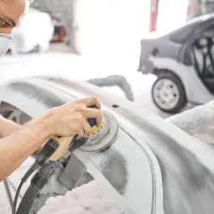 Pracownik w masce ochronnej wykonuje szlifowanie szpachli samochodowej na zderzaku. W tle widać auto w trakcie naprawy.