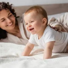 Un feliz bebe 7 meses gatea en la cama mientras su madre sonríe.
