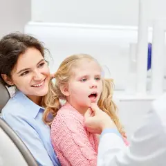 Uśmiechnięta mama towarzyszy córce podczas zabiegu lakowania zębów u dzieci. Dentysta bada jamę ustną dziewczynki.