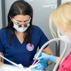 Dentystka w okularach powiększających i masce wykonuje zabieg. Asystentka trzyma ssak. Znieczulenie śródwięzadłowe zapewnia komfort pacjentowi.