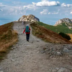 Turystka idzie ścieżką przez trawiaste zbocze w kierunku skalistych szczytów. W oddali widać malownicze **czerwone wierchy**.