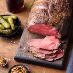 Un rôti de bœuf tranché, servi avec des cornichons, de la moutarde et des couverts sur une planche en bois.