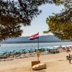 Chorwacka flaga powiewa nad pełną plażą z leżakami i parasolami. W tle błękitne morze i góry.