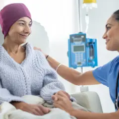 Eine Krankenschwester tröstet eine Patientin während des Chemotherapieablaufs. Die Patientin trägt eine lila Kopfbedeckung und eine Strickjacke.