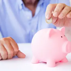 Mężczyzna wrzuca monetę do różowej świnki-skarbonki, zastanawiając się, czy lepsze będą lokaty czy konto oszczędnościowe.