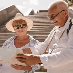 Uśmiechnięta para seniorów planuje wycieczki objazdowe samolotem z Gdańska, studiując mapę na tle schodów.