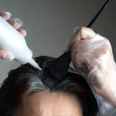 Ręce w rękawiczkach nakładają farbę na siwe odrosty u nasady ciemnych włosów.