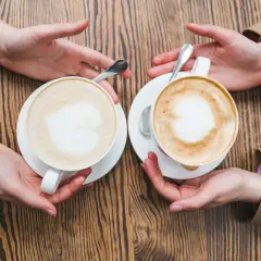 Zwei Cappuccinos mit Herz-Latte-Art, die Hände reichen sie sich über einen Holztisch. Wie viel Milch ist im perfekten Cappuccino?