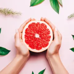 Dłonie trzymają połówkę grejpfruta, otoczone zielonymi liśćmi. Naturalne piękno i świeżość, jak wyciąg z pestek grejpfruta.