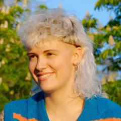 Ragazza con capelli biondi, corti sopra e lunghi dietro, stile anni '80, sorride all'aperto con alberi fioriti sullo sfondo.