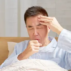 Mann im Bett mit geschlossenen Augen, der sich die Stirn reibt und hustet. Er leidet unter Kopfschmerzen beim Husten.