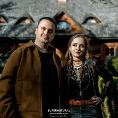 Para pozuje przed domem z kamiennym dachem. Mężczyzna w brązowej kurtce i kobieta w czarnej bluzce z wieloma naszyjnikami. Wyglądają jakby wyszli z planu filmu o straszydłach.