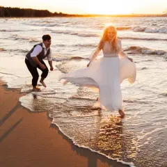 Szczęśliwa para podczas sesji na plaży o zachodzie słońca. Ona w zwiewnej sukni, on w eleganckim stroju, bawią się falami.