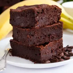 Stos puszystych brownie bananowych na talerzyku, obok dojrzały banan i ziarna kawy.