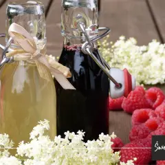 Dwa syropy z czarnego bzu w butelkach, otoczone kwiatami czarnego bzu i malinami. Syrop z czarnego bzu na przeziębienie i wzmocnienie odporności.