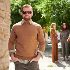 Mężczyzna w swetrze w kolorze camel i okularach przeciwsłonecznych stoi na tle kamiennej ściany i zieleni.