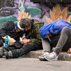 Trzech mężczyzn siedzi na chodniku, zgarbionych, z butelką napoju obok. Obraz ten może symbolizować problem uzależnień, w tym fentanyl w Polsce.