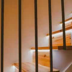 Drewniane stopnie schodów z subtelnym oświetleniem LED w ścianie. Czarne, pionowe balustrady tworzą nowoczesny kontrast.
