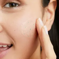 En kvinna applicerar en ansiktsskrubb med små, vita pormaskar-liknande korn för att exfoliera huden.