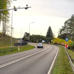 Znak informuje o systemie radarowym pomiaru prędkości. Na drodze widać samochód i kamery nad drogą.