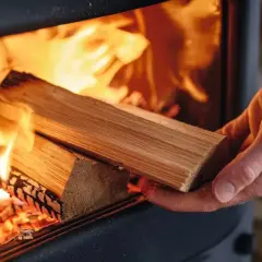 Eine Hand legt Holz in einen brennenden Kaminofen. Die Flammen lodern hell und wärmend.