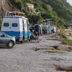 Une rangée de camping-cars en Grèce, certains garés sur un chemin de gravier près d'une plage. Des gens se détendent à l'extérieur.