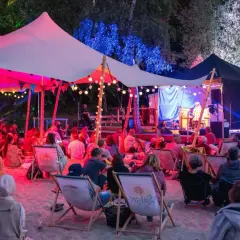 Menschen genießen ein **musik und stille festival** unter einem beleuchteten Zelt. Musiker spielen auf einer Bühne, während das Publikum auf Liegestühlen im Sand sitzt.