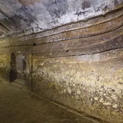 Interno di una domus de janas s'incantu, con pareti scavate e gradini che conducono a un'apertura.