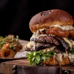 Un généreux burger maison avec plusieurs steaks, fromage fondu et laitue fraîche, servi sur une planche en bois.