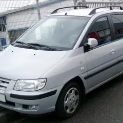 Srebrny Hyundai Matrix stoi przed budynkiem z niebieskimi drzwiami i metalowym ogrodzeniem.