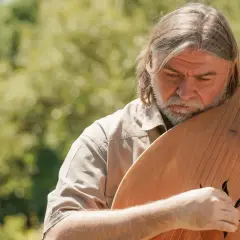 Mężczyzna z brodą i długimi włosami gra na bandurze na tle zieleni.