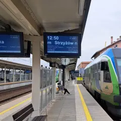 Na peronie kolejowym widać tablice informacyjne z rozkładem jazdy, w tym wskaźnik w4 pokazujący odjazd pociągu do Medyki. Obok stoi zielono-niebieski pociąg.