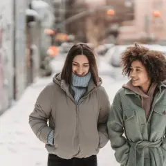 Deux amies discutent en marchant dans la neige. Une idée outfit hiver parfaite : doudoune beige et manteau vert ceinturé.