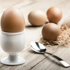 Jajko na miękko w kieliszku, obok łyżeczka i kilka jajek na drewnianym tle.