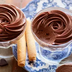 Due coppette di mousse al cioccolato ricetta classica, guarnite con rotoli di wafer e cuoricini di cioccolato.