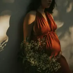Kobieta w pomarańczowej sukience, z dłońmi na brzuchu, jakby we śnie o ciąży. W tle cień liści.