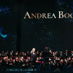 Andrea Bocelli koncert z orkiestrą symfoniczną. Wokalista w garniturze stoi na scenie, dyrygent kieruje muzykami.