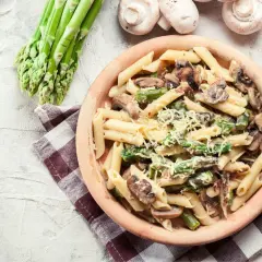 Penne z kremowym sosem, szparagami i pieczarkami, posypane parmezanem. Obok pęczek świeżych szparagów i kilka pieczarek.