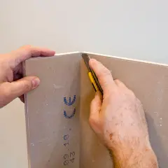Mani che tengono un cartongesso mentre un taglierino viene usato per tagliare il materiale. Impara come tagliare il cartongesso con precisione.