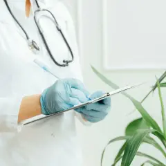Medico con camice bianco e guanti blu scrive su un blocco appunti. Lo stetoscopio è appeso al collo. Il significato di "extramoenia" è legato a ciò che è fuori dalle mura, come un consulto medico fuori dall'ospedale.