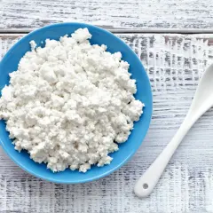 Serek wiejski w niebieskiej miseczce obok białej łyżki na drewnianym tle.