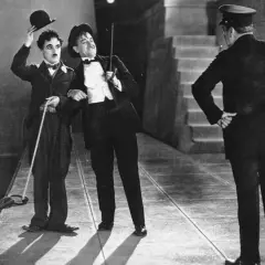 Charlie Chaplin i jego towarzysz w eleganckich strojach, z kapeluszami i laskami, w scenie z okresu kina niemego. Policjant stoi tyłem.