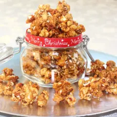 En burk fylld med karamelliserade popcorn, dekorerad med ett rött band med texten "Merry Christmas". Några popcorn ligger utspridda på ett fat.