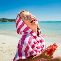 Niña riendo en la playa con una toalla a rayas y sandía, perfecta comida para llevar a la playa con niños.