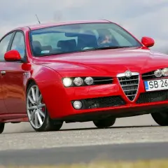 Czerwone Alfa Romeo 159 1.9 JTD na drodze, z kierowcą za kierownicą.