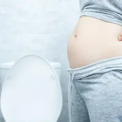 Frau mit Blähbauch hält sich den Bauch. Was hilft gegen Blähbauch? Die Toilette im Hintergrund deutet auf Verdauungsprobleme hin.